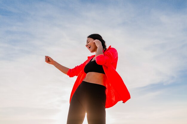 Young smiling attractive slim woman doing sports in morning sunrise dancing on sea beach in sports wear, healthy lifestyle, listening to music on earphones, wearing pink windbreaker jacket, having fun