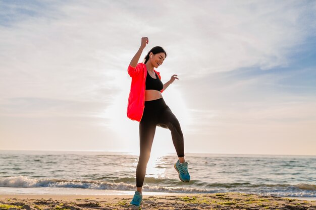 Young smiling attractive slim woman doing sports in morning sunrise jumping on sea beach in sports wear, healthy lifestyle, listening to music on earphones, wearing pink windbreaker jacket, having fun