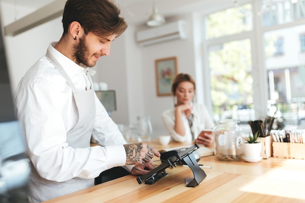 Young smiling barista in apron and white shirt using cash counter in coffee shop while thoughtful girl on background sitting at the counter with mobile phone Cool boy working as barista at cafe