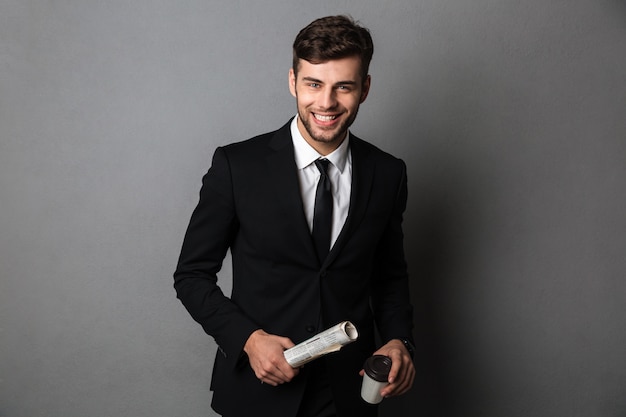 Young smiling bearded man in formalwear holding newspaper and cup of coffee, 