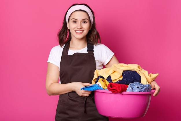 ピンクの盆地で汚れた服のアイテムでいっぱいに歩いている若い笑顔の美しい女性は、両手でそれを持っていると、ポジティブに見えます。忙しい魅力的な主婦スタンドに孤立したピンク。