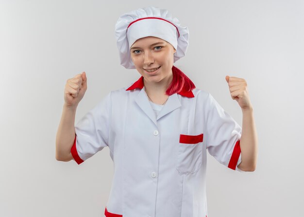 Young smiling blonde female chef in chef uniform holds fists up isolated on white wall