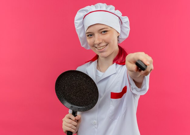 요리사 제복을 입은 젊은 웃는 금발 여성 요리사는 프라이팬을 보유하고 분홍색 벽에 고립 된 칼을 보유하고 있습니다.