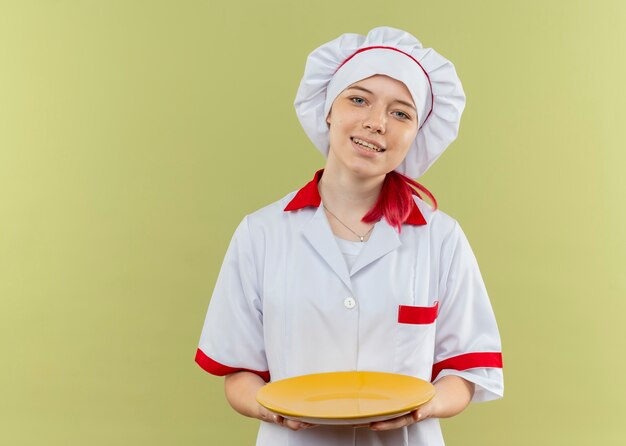 Young smiling blonde female chef in chef uniform holds plate and looks isolated on green wall