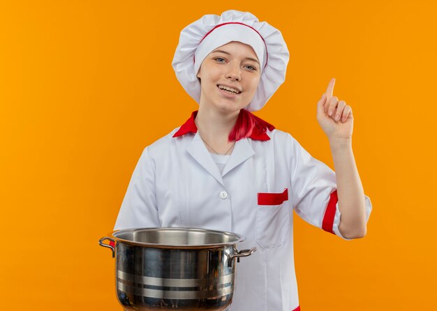요리사 유니폼에 젊은 웃는 금발 여성 요리사 냄비를 보유하고 오렌지 벽에 고립 된 포인트