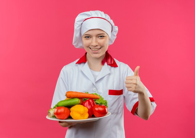 Young smiling blonde female chef in chef uniform holds vegetables on plate and thumbs up isolated on pink wall