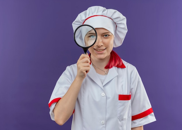 Бесплатное фото Молодая улыбающаяся блондинка-шеф-повар в униформе смотрит через увеличительное стекло или лупу, изолированную на фиолетовой стене