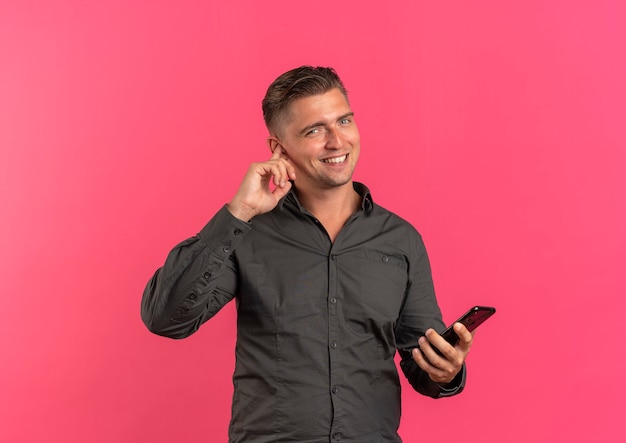 Young smiling blonde handsome man closes ear with finger and holds phone isolated on pink background with copy space