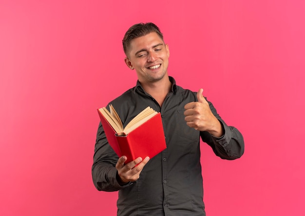 Молодой улыбающийся блондин красивый мужчина держит книгу и пальцы вверх изолированы на розовом фоне с копией пространства