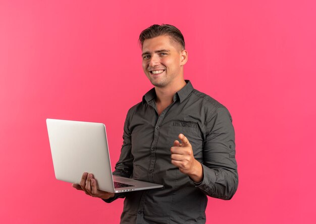 Young smiling blonde handsome man holds laptop and points at camera isolated on pink background with copy space