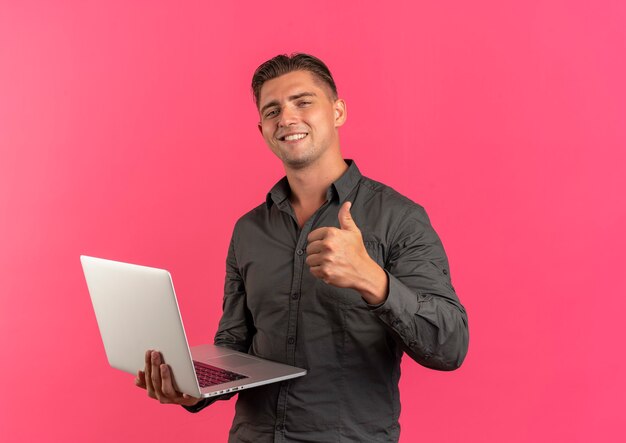 Young smiling blonde handsome man holds laptop and thumbs up isolated on pink background with copy space