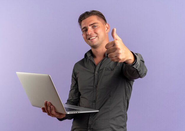 Young smiling blonde handsome man holds laptop and thumbs up looking at camera isolated on violet background with copy space