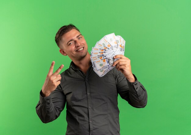 Young smiling blonde handsome man holds money and gestures victory hand sign isolated on green space with copy space