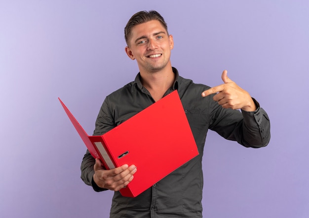 Young smiling blonde handsome man holds and points at file folder isolated on violet space with copy space