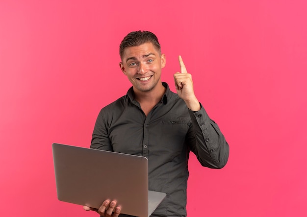 Young smiling blonde handsome manholds laptop and points up looking at camera isolated on pink background with copy space