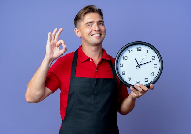 Young smiling blonde male barber in uniform holds clock gesturing ok hand sign isolated on violet space with copy space
