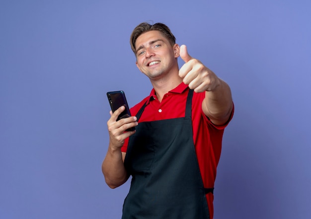 young smiling blonde male barber in uniform holds phone thumbs up isolated on violet background