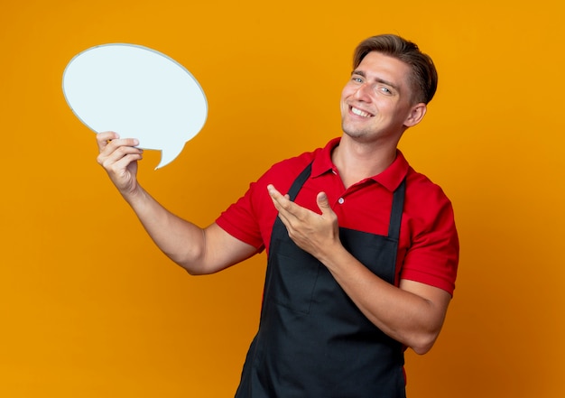 Young smiling blonde male barber in uniform holds and points at chat bubble isolated on orange space with copy space