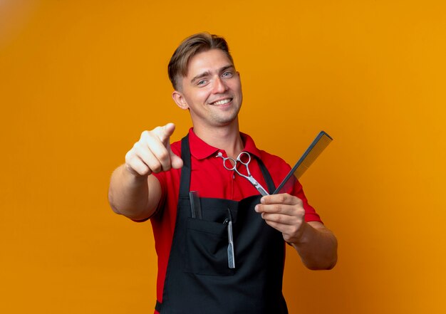 Молодой улыбающийся белокурый мужчина-парикмахер в униформе указывает на камеру, держащую ножницы и расческу, изолированную на оранжевом пространстве с копией пространства