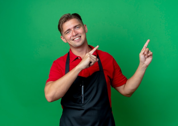 Молодой улыбающийся белокурый мужчина-парикмахер в униформе указывает сбоку двумя руками, изолированными на зеленом пространстве с копией пространства