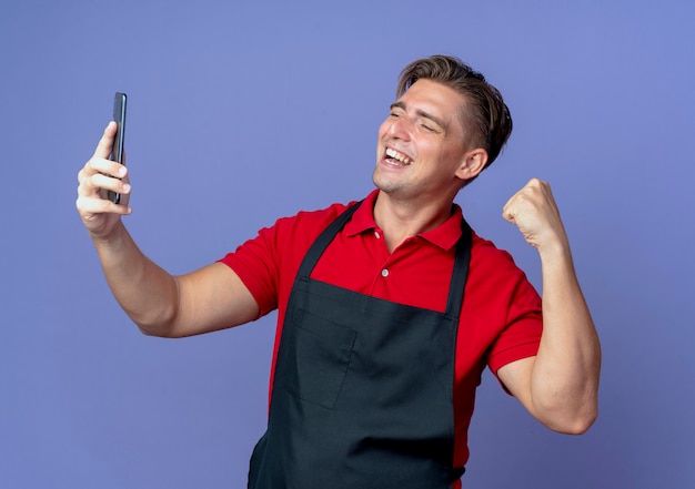 молодой улыбающийся блондин мужчина-парикмахер в униформе поднимает кулак, глядя на телефон