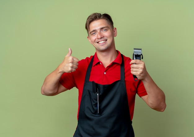 Il giovane barbiere maschio biondo sorridente in uniforme sfoglia e tiene il tagliatore di capelli isolato sullo spazio verde oliva con lo spazio della copia