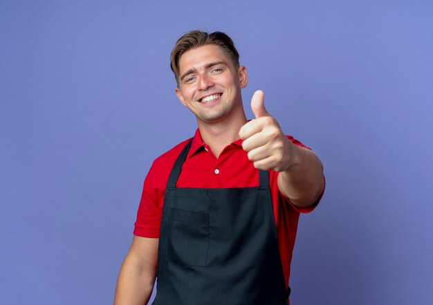 Young smiling blonde male barber in uniform thumbs up isolated on violet space with copy space