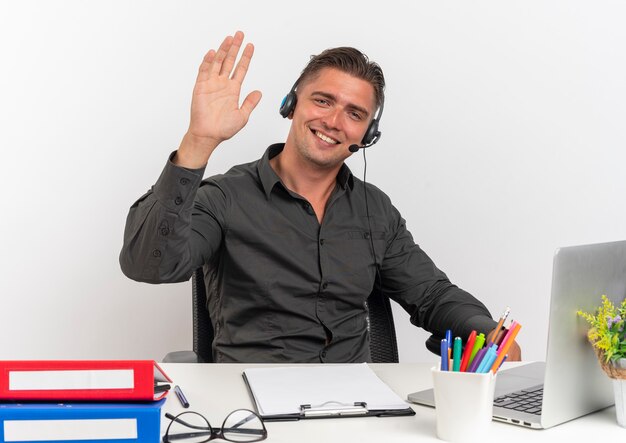 Молодой улыбающийся белокурый человек офисного работника в наушниках сидит за столом с офисными инструментами, используя ноутбук, смотрит в камеру с поднятой рукой, изолированной на белом фоне с копией пространства