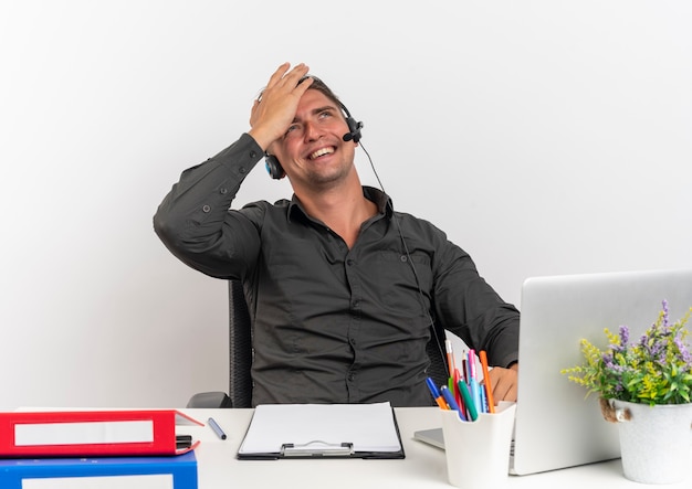 Young smiling blonde office worker man on headphones sits at desk with office tools using laptop puts hand on head looking up isolated on white background with copy space