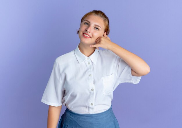 Young smiling blonde russian girl gestures call me sign isolated on purple background with copy space