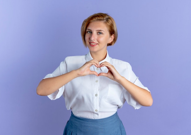 Молодая улыбающаяся русская блондинка жестикулирует знак руки сердца, изолированные на фиолетовом фоне с копией пространства
