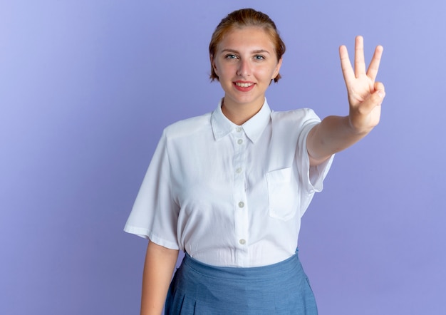 young smiling blonde russian girl gestures three isolated on purple background with copy space