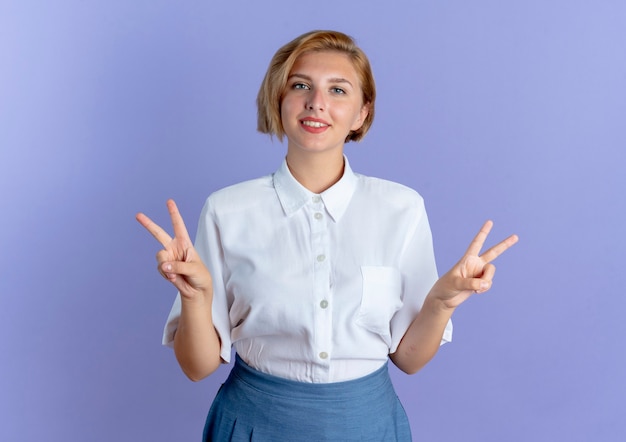 La giovane ragazza russa bionda sorridente gesticola il segno della mano di vittoria con entrambe le mani isolate su fondo viola con lo spazio della copia