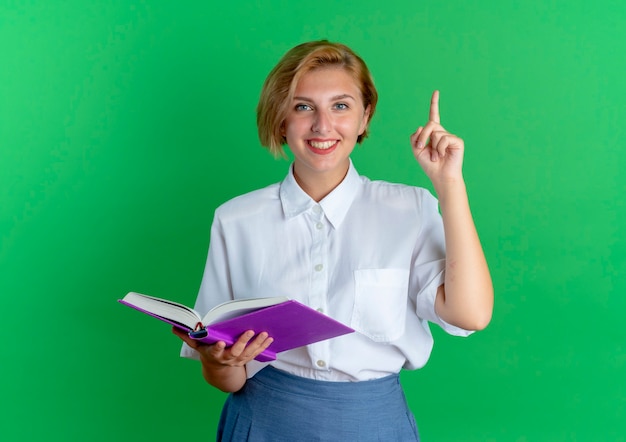 Бесплатное фото Молодая улыбающаяся русская блондинка держит книгу, направленную вверх, изолированную на зеленом фоне с копией пространства