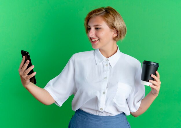 Young smiling blonde russian girl holds coffee cup looking at phone isolated on green background with copy space