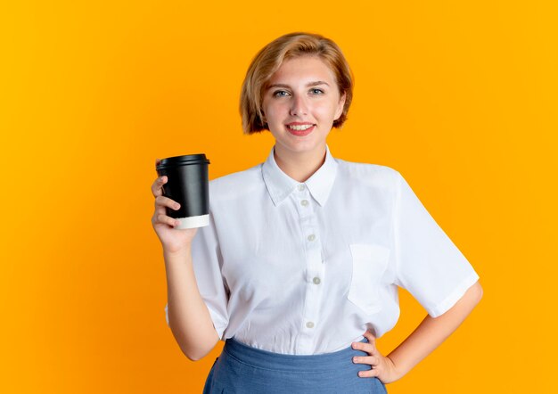 젊은 웃는 금발 러시아 여자 보유 커피 컵 복사 공간이 오렌지 배경에 고립 허리에 손을 둔다
