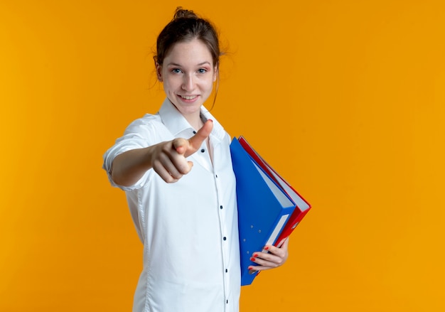 Free photo young smiling blonde russian girl holds file folders pointing at camera isolated on orange space with copy space