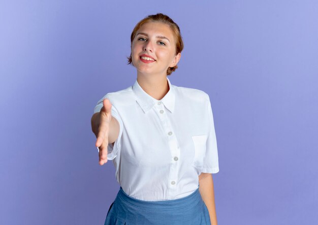 Young smiling blonde russian girl holds hand out isolated on purple background with copy space