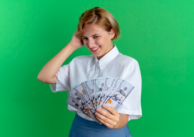 Free photo young smiling blonde russian girl holds and looks at money puts hand on head isolated on green background with copy space