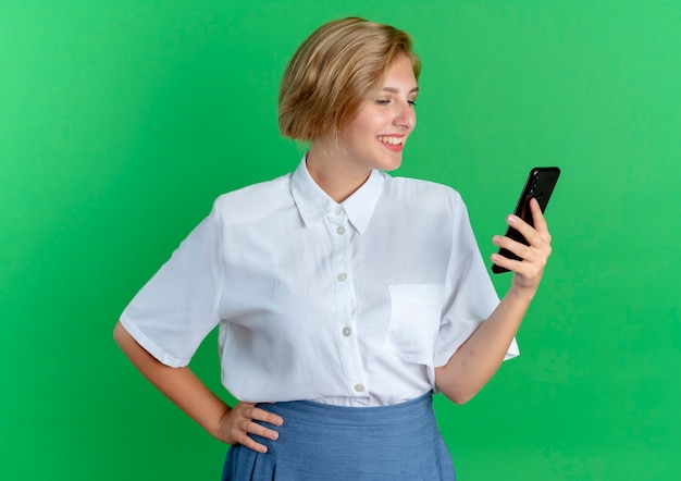 Young smiling blonde russian girl holds and looks at phone isolated on green background with copy space