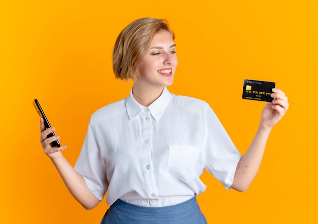 若い笑顔の金髪のロシアの女の子が電話を持ってクレジットカードを見る