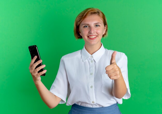 Young smiling blonde russian girl holds phone thumbs up isolated on green background with copy space