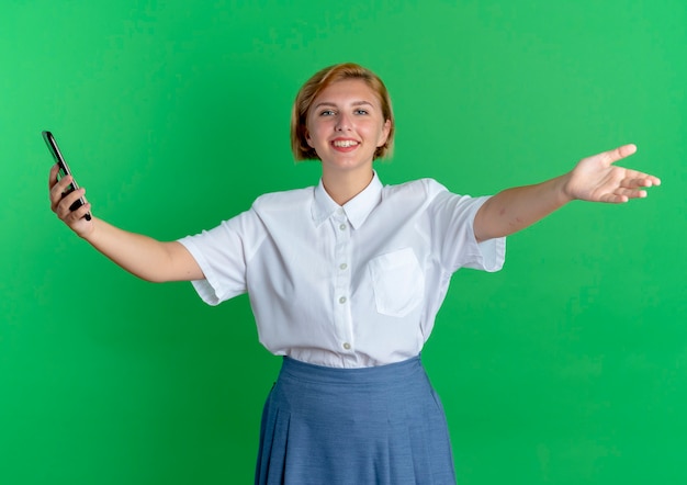 若い笑顔のブロンドのロシアの女の子は、コピースペースで緑の背景に分離された両手を広げて電話を保持します。