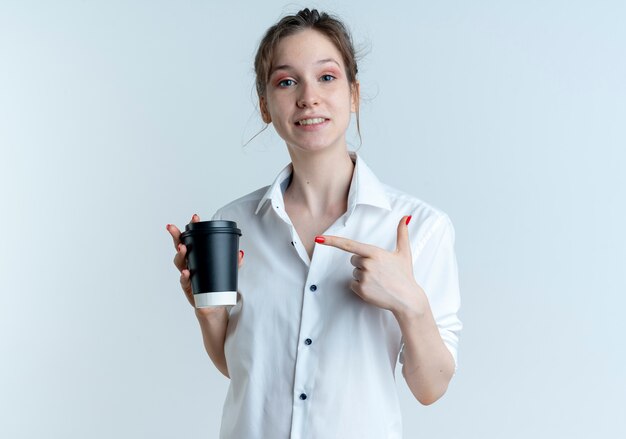 Young smiling blonde russian girl holds and points at coffee cup isolated on white space with copy space