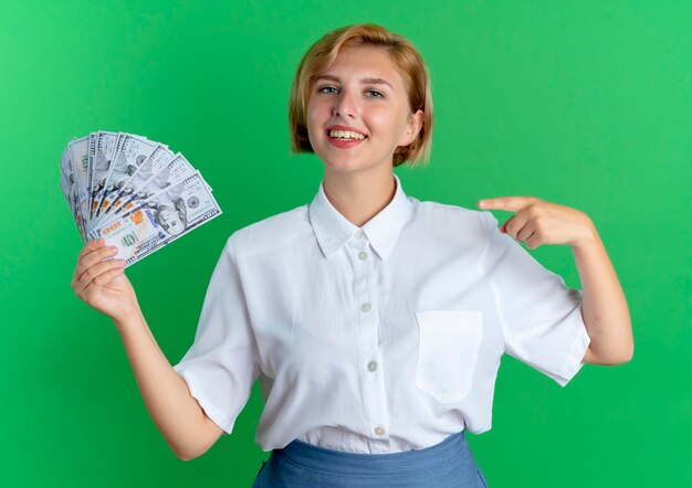 Молодая улыбающаяся белокурая русская девушка держит и указывает на деньги, изолированные на зеленом пространстве с копией пространства
