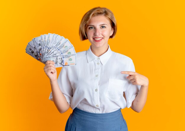 Young smiling blonde russian girl holds and points at money isolated on orange background with copy space