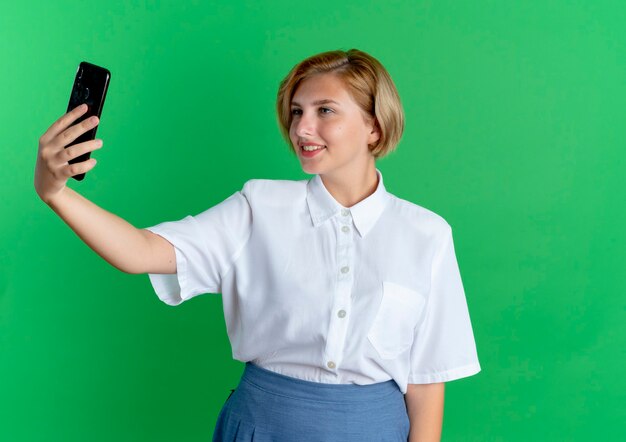 Young smiling blonde russian girl looks at phone isolated on green background with copy space