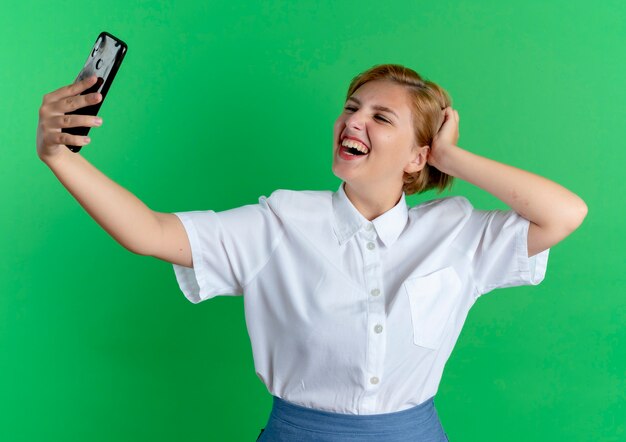 young smiling blonde russian girl looks at phone taking selfie puts hand on head