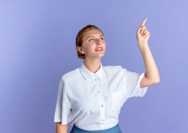 Young smiling blonde russian girl looks and points at side isolated on purple background with copy space