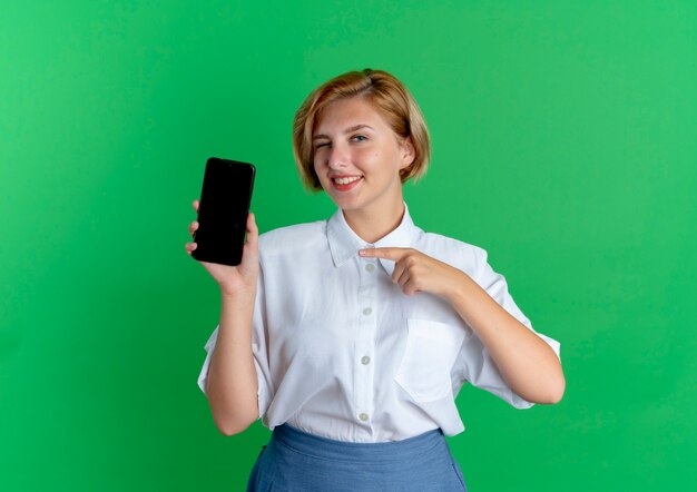 young smiling blonde russian girl points at phone isolated on green background with copy space